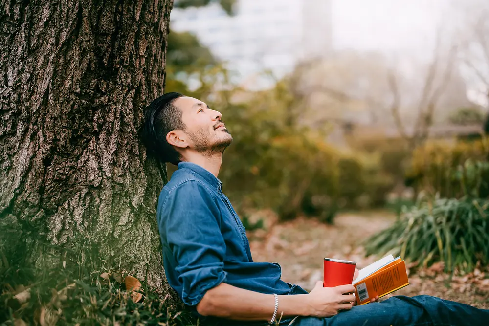 Un hombre con un libro recostado en un árbol.